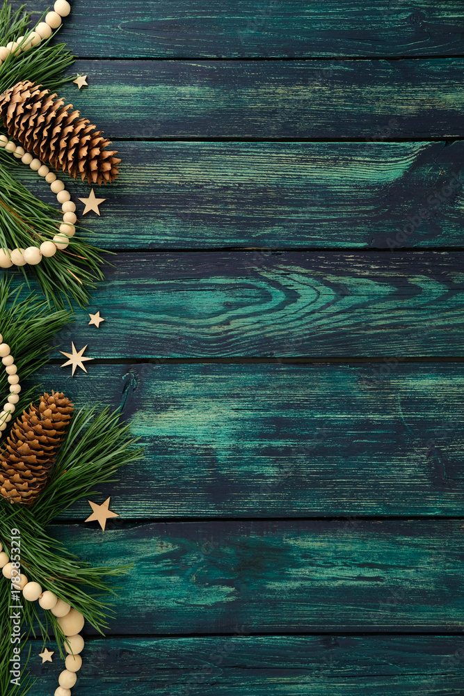 Obraz premium Rustic Christmas border with pine branches, pinecones, wooden beads, and stars on dark teal wooden background, festive frame with copy space.