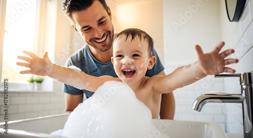 Father smiles as his young child splashes in a bubble bath, arms outstretched. Bathroom setting, natural light. Happy family moment.
