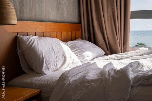 Unmade bed with white pillows and duvet in sunny hotel room with sea view