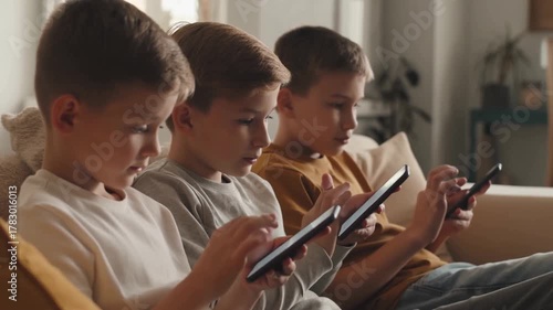 Three young boys are engrossed in using their devices, each focused on their respective screens. They are sitting together in a comfortable indoor setting. Stock Video
