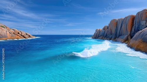 Vibrant Blue Ocean Waves Crash Against Rugged Rocky Cliffs Under a Clear Sunny Sky