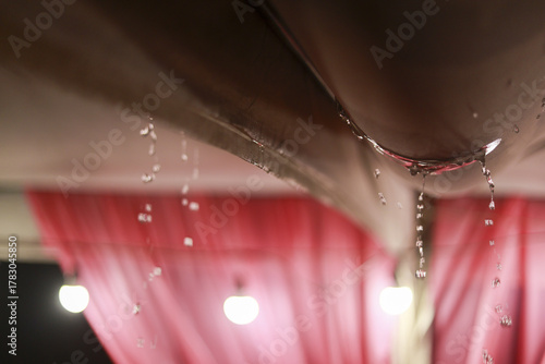 Close up view of concerning water drop dripping from damaged ceiling. serious indoor leak and plumbing problem causing water damage in home with pink background