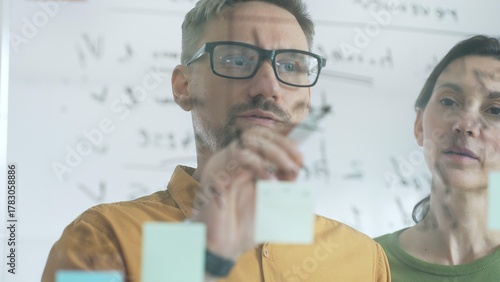 Two business people colleagues are analyzing notes on a transparent whiteboard during a brainstorming session, working together to develop innovative solutions and strategies for their deal
