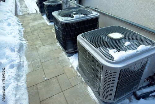 Outdoor HVAC units in winter snow.Several outdoor air conditioning and heat pump units covered with snow near a building wall, showing winter conditions and energy system maintenance.
