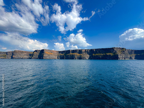 Majestic sea cliffs rise above calm blue ocean beneath a bright sky with scattered clouds on a sunny day
