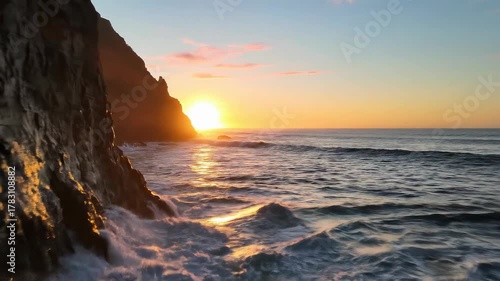 Golden sunset over the ocean with waves crashing against a rocky cliffside.