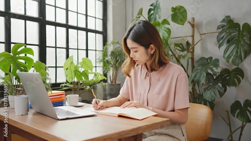 Creative Workspace with Study: A young individual engages in focused work, pen in hand, surrounded by the inviting ambiance of a modern home office with laptop and nature-inspired décor. 