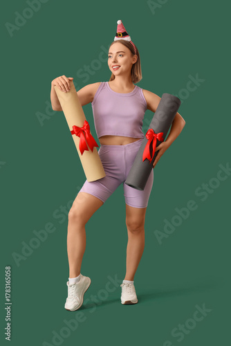 Sporty young woman in elf hat with yoga mats as Christmas gifts on green back...