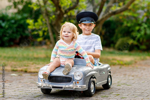 Two happy children playing with big old toy car in summer garden, outdoors. Kid boy driving car with little toddler girl inside. Laughing and smiling kids. Family, childhood, lifestyle concept