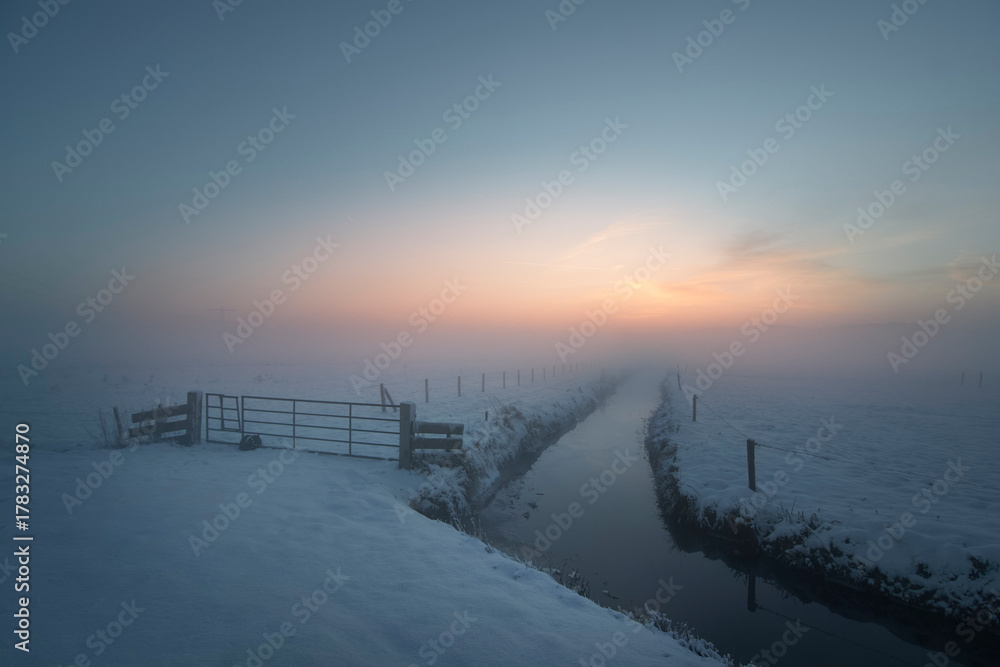 Fototapeta premium Snowy rural landscape with fog and frozen canal at winter sunrise