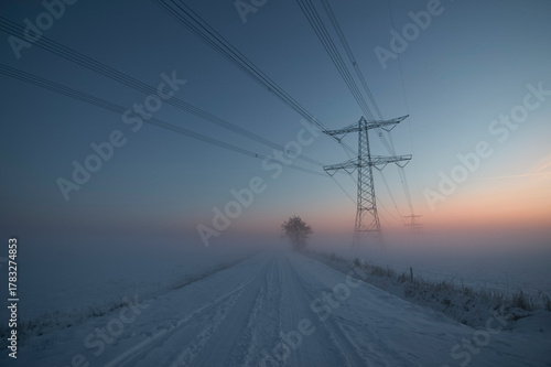 Snowy winter road with high voltage power line pylon fading into misty sunrise, rural landscape