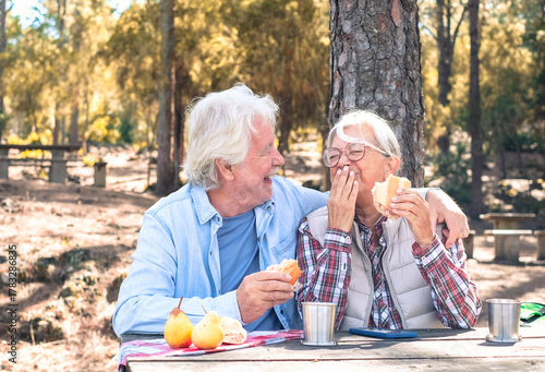 Happy senior couple enjoying a picnic outdoors in a sunny forest. The elderly man and woman are sitting at a wooden table, laughing sharing food and drinks. Positive retired lifestyle in nature