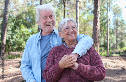 Happy senior couple enjoying a moment of affection and emotion outdoors in a sunny forest. The elderly man hugs his partner while both smile warmly. Healthy retirement lifestyle in nature