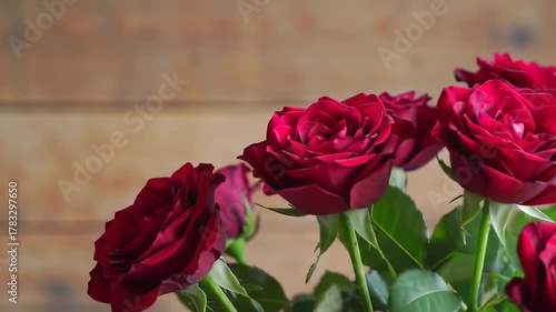 A close-up shot of several deep red roses in full bloom with green leaves. A wooden surface background
