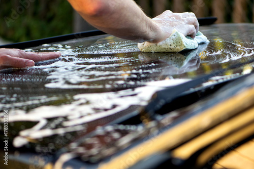 a hand with a washcloth and soap suds washes the hood of the car. Man washing car hood with washcloth and shampoo, auto covered with foam, car wash service. cleaning the black surface