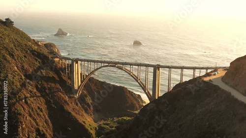 A slow aerial pan captures the iconic Bixby Creek Bridge on California's coast at golden hour, showing its grand arch over a canyon by the ocean.