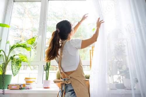 Woman in apron hangs transparent tulle curtains on large windows in the house inside the interior with potted plants. Spring cleaning, tidying up