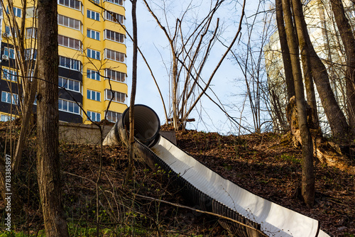 Large corrugated storm drain pipe descends steep wooded slope. Urban development meets nature