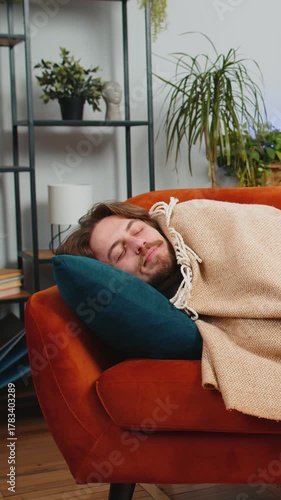 Tired young man lying down in bed taking a rest at home apartment room. Carefree Caucasian guy napping, falling asleep on comfortable orange sofa with pillows. Closed his eyes enjoy daytime nap alone