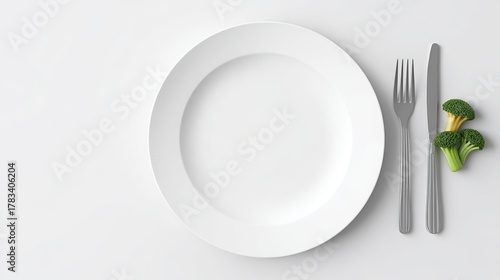 Top down view of an empty white dinner plate silver fork knife and fresh green broccoli florets on a clean white surface