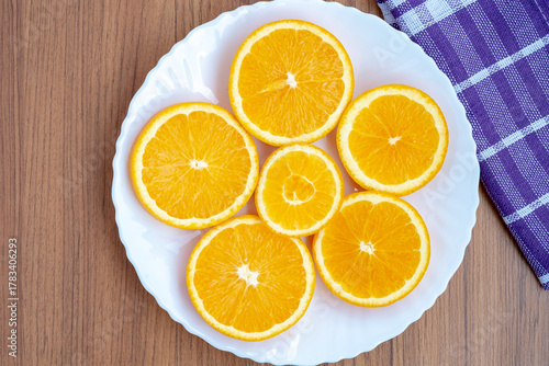 A top-down view shows several round slices of orange fruit (Citrus × sinensis) arranged on a white plate. The plate rests on a wooden surface next to a purple checkered cloth.