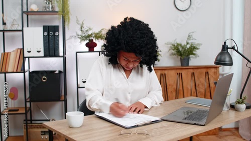 African American businesswoman at home office reviews documents spots error crumples paper frustrated tossing it aside. Freelancer girl at table sighs upset plans correction before submitting file up.