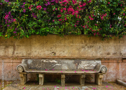 An elegant Neoclassical stone bench with Bougainvillea flowers in Palermo, Italy. Located on Mure delle Cattive in Kalsa district. The marble bench features scrolled volute armrests & legs with 