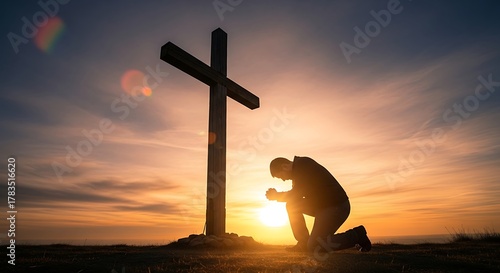 Silhouette of a man praying at the foot of the cross at sunset.