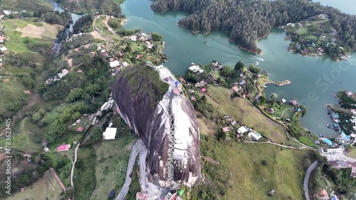A dynamic aerial pulls back from the top of the Guatapé rock and pans upward, revealing the entire massive monolith, surrounding lake, islands, and landscape under a cloudy sky