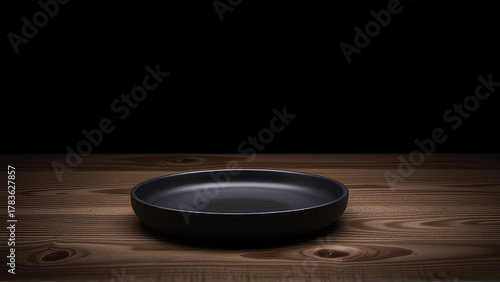 Black empty plate on wooden table with dark background, minimalistic kitchenware scene with soft lighting and natural wood texture, cozy and calm atmosphere