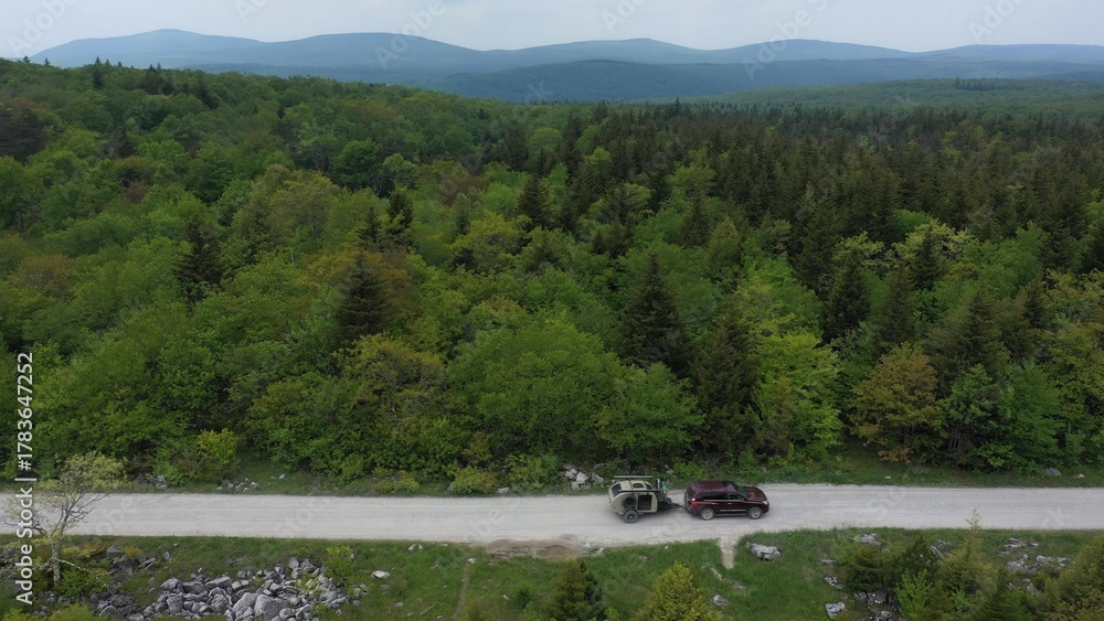 Naklejka premium A family enjoys a camping adventure in a teardrop trailer, parked along a dirt road surrounded by vibrant green trees under a cloudy sky.