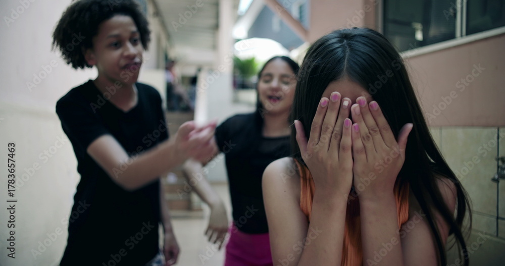 Obraz premium Teenager covering face in distress while friends laugh in suburban alleyway, representing youth bullying, peer relationships, and emotional challenges