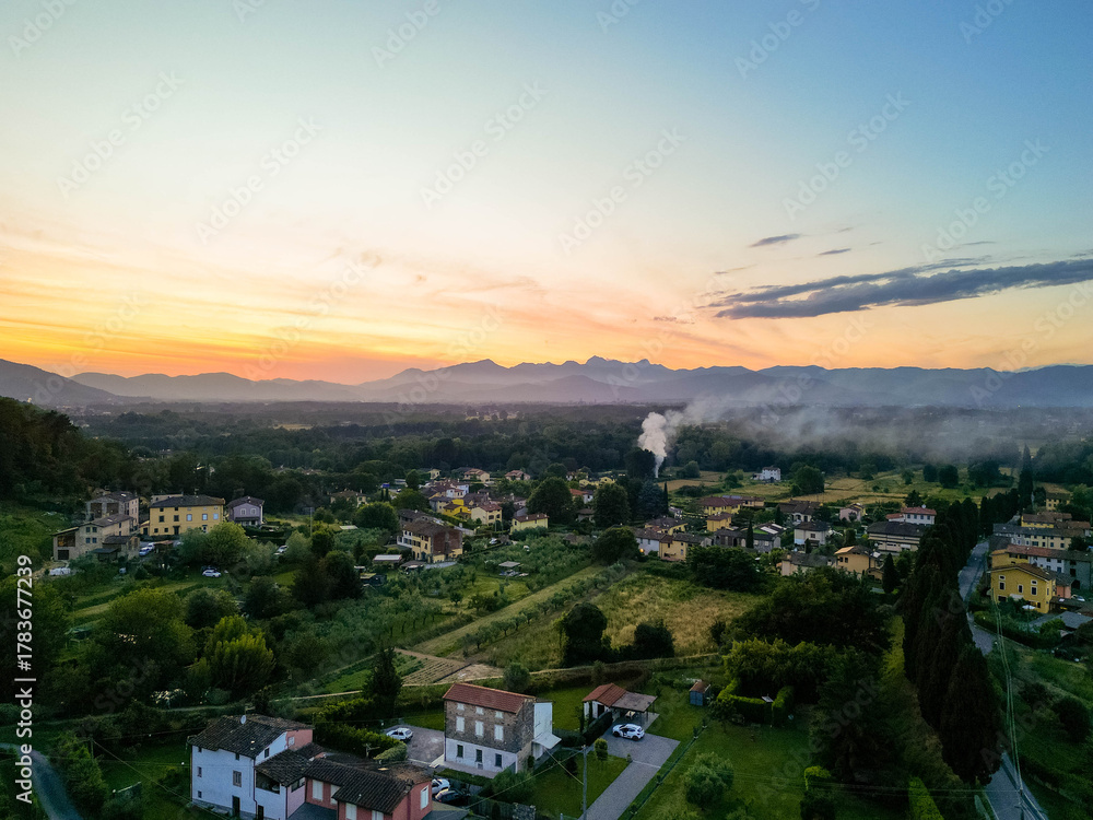 Fototapeta premium Aerial view of the Tuscan countryside during sunset, showing rustic village houses surrounded by olive groves, cypress trees, and rolling hills.
