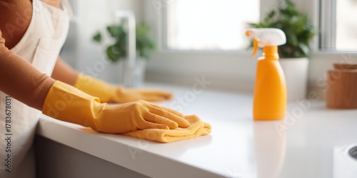Fototapeta Naklejka Na Ścianę i Meble -  Woman in yellow gloves wiping a white kitchen counter with an orange microfiber cloth, a spray bottle beside her. Home cleanliness concept.