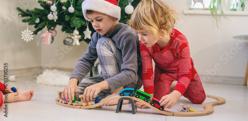 A boy and a girl are playing with a wooden toy train near a Christmas tree at home.