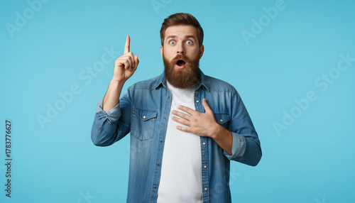 Bearded man in casual shirt pointing up with surprised face, solid cyan blue background.