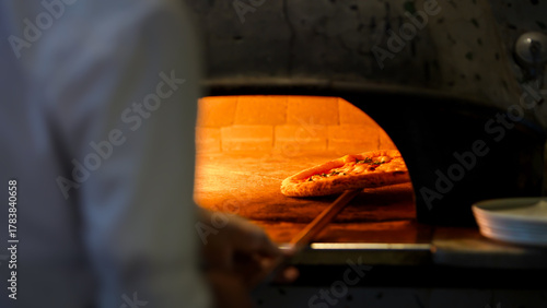 A pizza rests on a stone in a wood fired oven, with the fire heat cooking