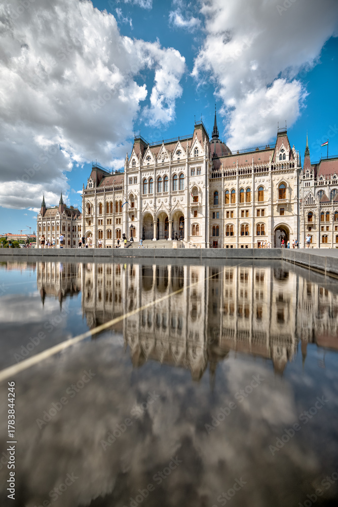 Naklejka premium Budapest, Hungary - July 27, 2025: Reflection of the Hungarian Parliament Building in the shallow pool at Kossuth Lajos Square under a dramatic summer sky