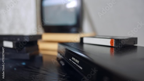 VHS player, several tapes and shimmering screen. A cassette appears from the player and hand puts it on the pile.