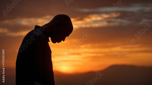 A person stands with a bowed head against a vibrant sunset background. The warm colors of the sky create a peaceful atmosphere, encouraging moments of reflection and tranquility.