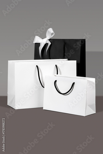 Three paper shopping bags, white and black with handles on grey background. Concept of premium gifting, stylish packaging, holiday shopping display and elegant branding for festive occasions. Vertical
