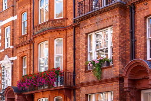 Fototapeta Naklejka Na Ścianę i Meble -  Red brick facade of a British-style building with a small balcony decorated by vibrant flowers.