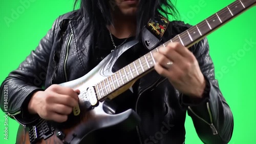 Dynamic closeup of a rock guitarist with long dark hair and a leather jacket passionately playing an electric guitar against a vibrant green screen showcasing intense musical performance and energeti.