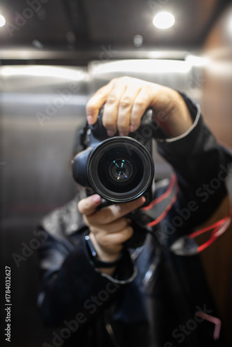 Photographer taking a selfie or video with a dslr camera held up close, standing in a mirrored elevator with stainless steel walls