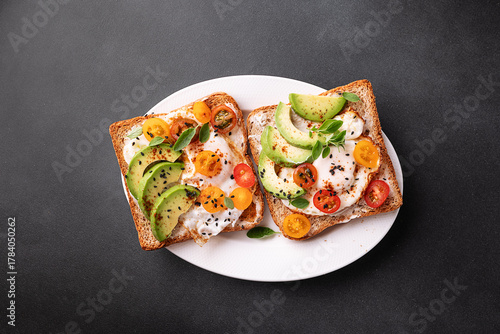 Tasty homemade toast for breakfast with fried eggs, cream cheese, avocado, cherry tomatoes and fresh oregano leaves on black stone background top view.