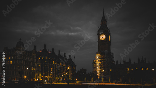 Night view of Big Ben and Westminster under brooding clouds, with warm clock glow and city lights streaking across the frame.