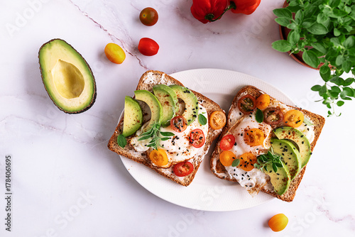 Tasty homemade toasts for breakfast with fried eggs, cream cheese, avocado, cherry tomatoes and fresh oregano leaves on white marble background top view.