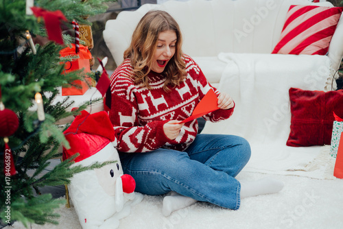 Young 30s woman getting Christmas greeting card from her beloved on background of xmas tree at cozy living room with festive interior. Surprised lady pleasure reading letter at home.