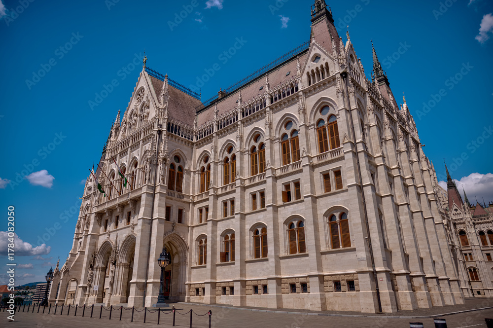 Fototapeta premium Budapest, Hungary - July 27, 2025: The Hungarian Parliament Building viewed from its garden side, showcasing its magnificent Neo-Gothic architecture with detailed spires and ornate stonework.
