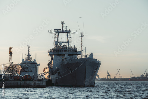 Telephoto view of large naval ships docked at industrial harbor during sunset, with cranes and calm sea creating dramatic maritime atmosphere and strong steel silhouettes